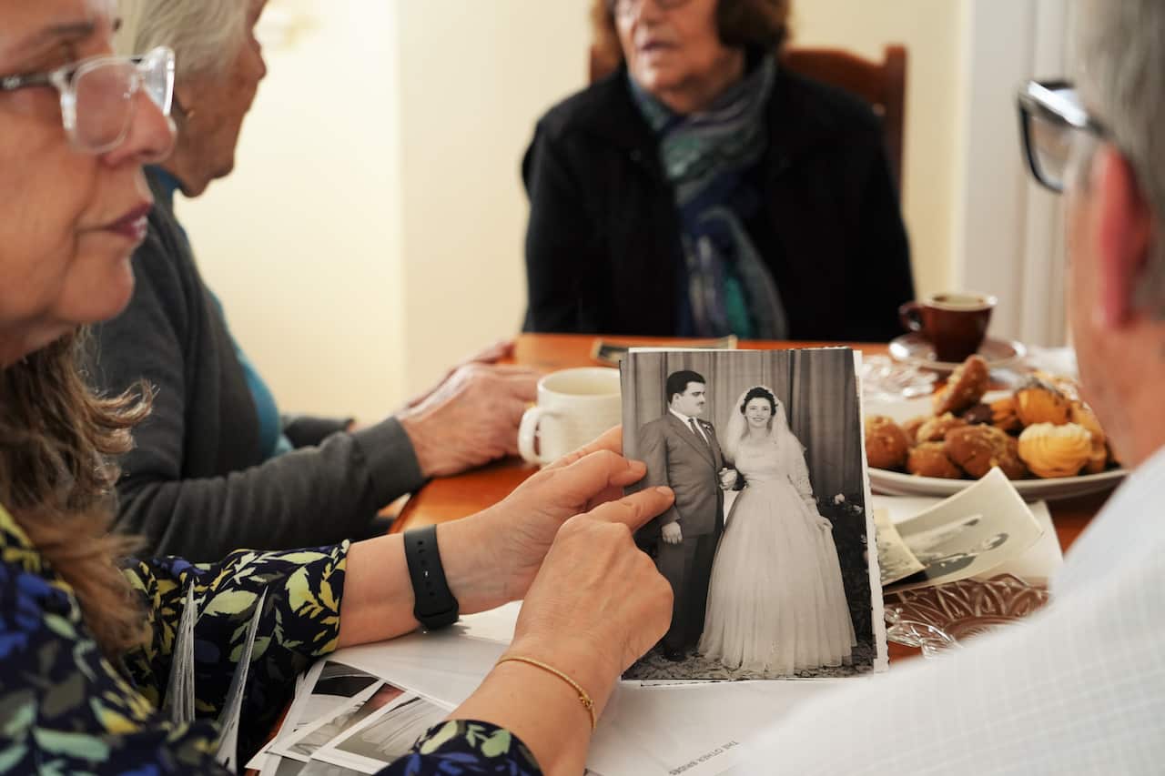 A woman wearing glasses holds a black-and-white photo of a young couple on their wedding day. In the background, two older women talk to each other, and a man stands off to their right. 