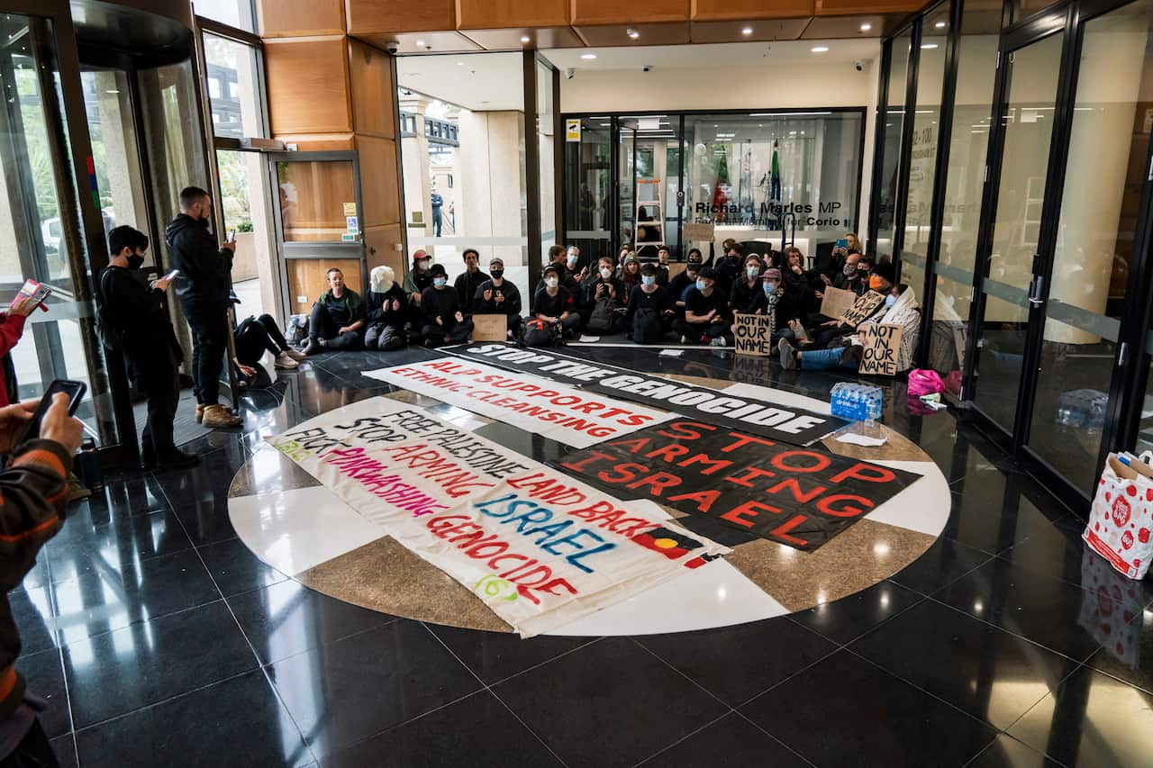 Protesters seated on the ground in the foyer of a building. In front of them are large signs.