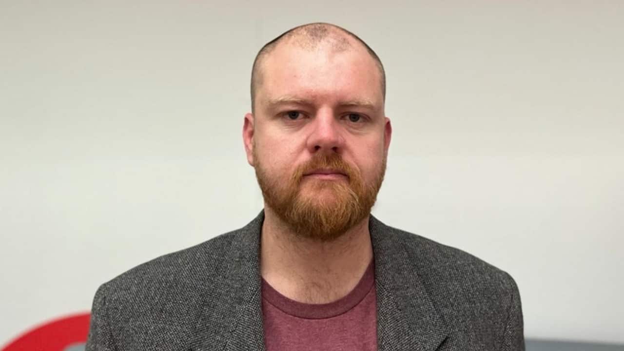 a middle aged man with a beard stands in an office