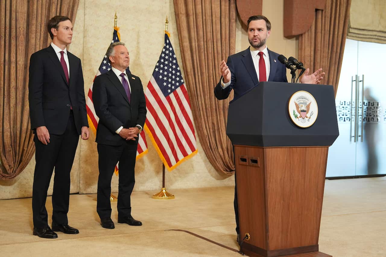 A white man in a suit, with brown hair and a beard, speaking at a podium. Two other men stand to his side, American flags hang on a flagpole. 