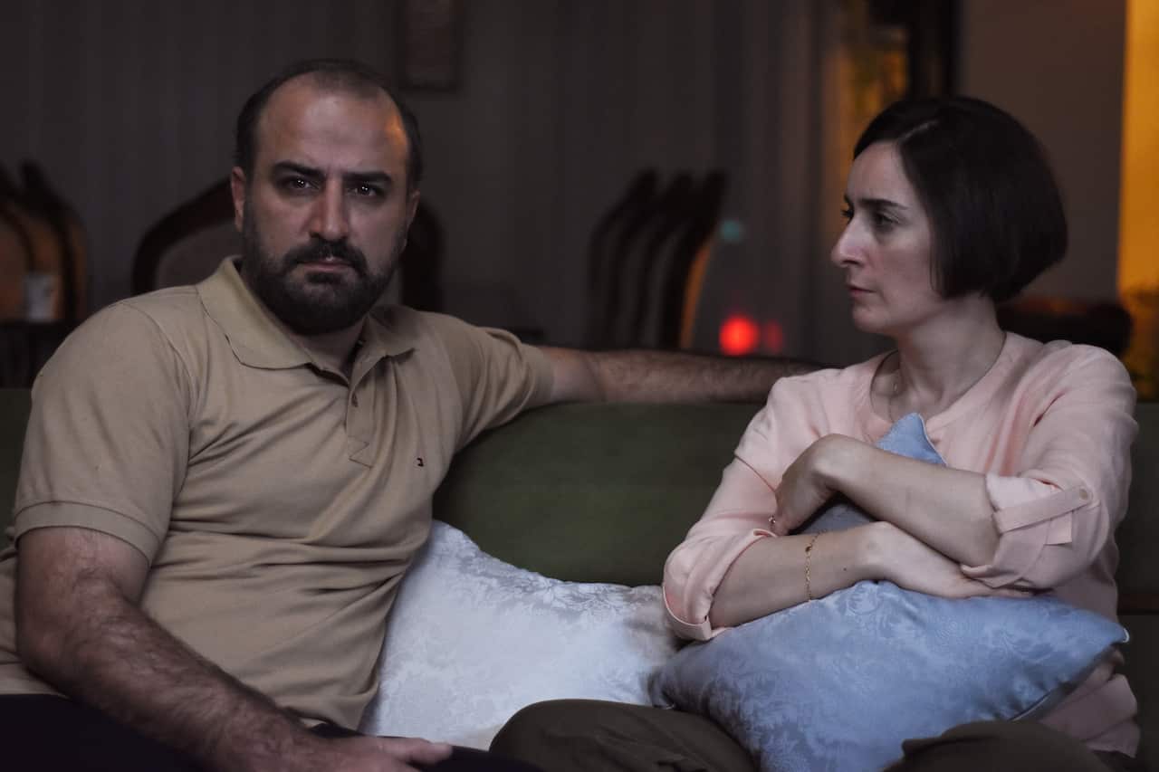 A woman and a man on a couch. The man, who has a beard and is wearing a light brown polo shirt, is looking straight forward. The woman is holding a blue pillow and looking at him.
