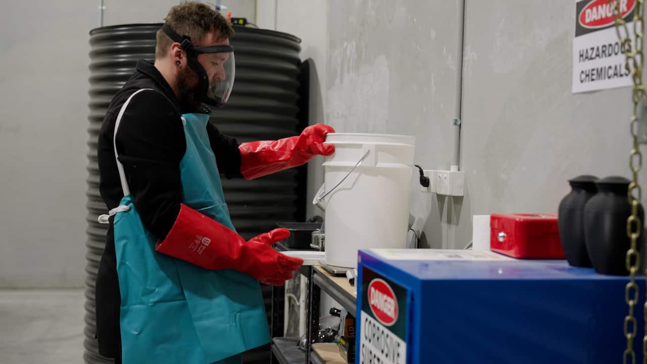 A man wearing a green apron, large red rubber gloves and a perspex face mask holding a bucket. 