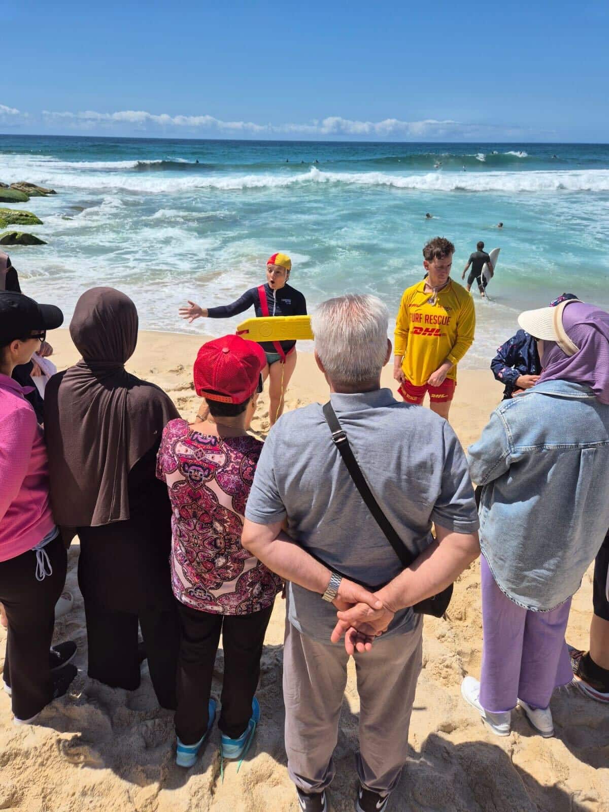 Men and women, including a woman in headscarf and a woman in a red cap, watch two life savers in yellow uniforms demonstrating a life saving technique before a choppy blue ocean.