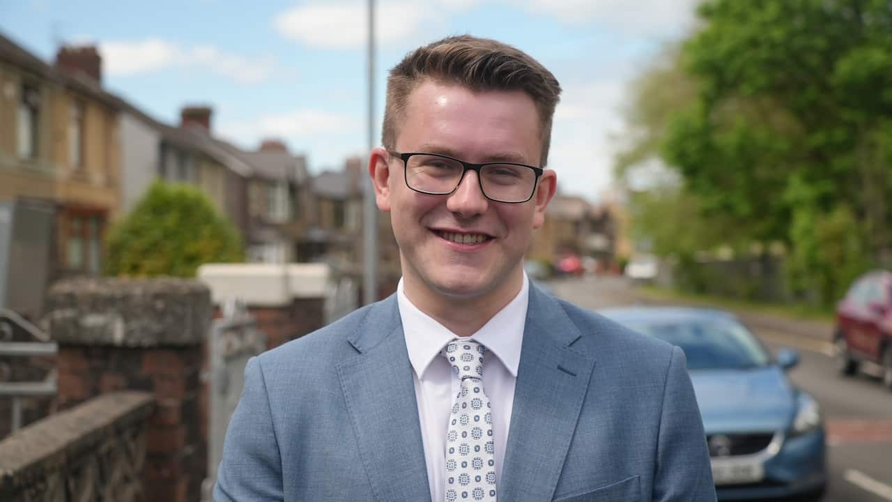 A smiling man with glasses, wearing a blue suit and patterned tie, stands on a residential street with houses and cars blurred in the background.