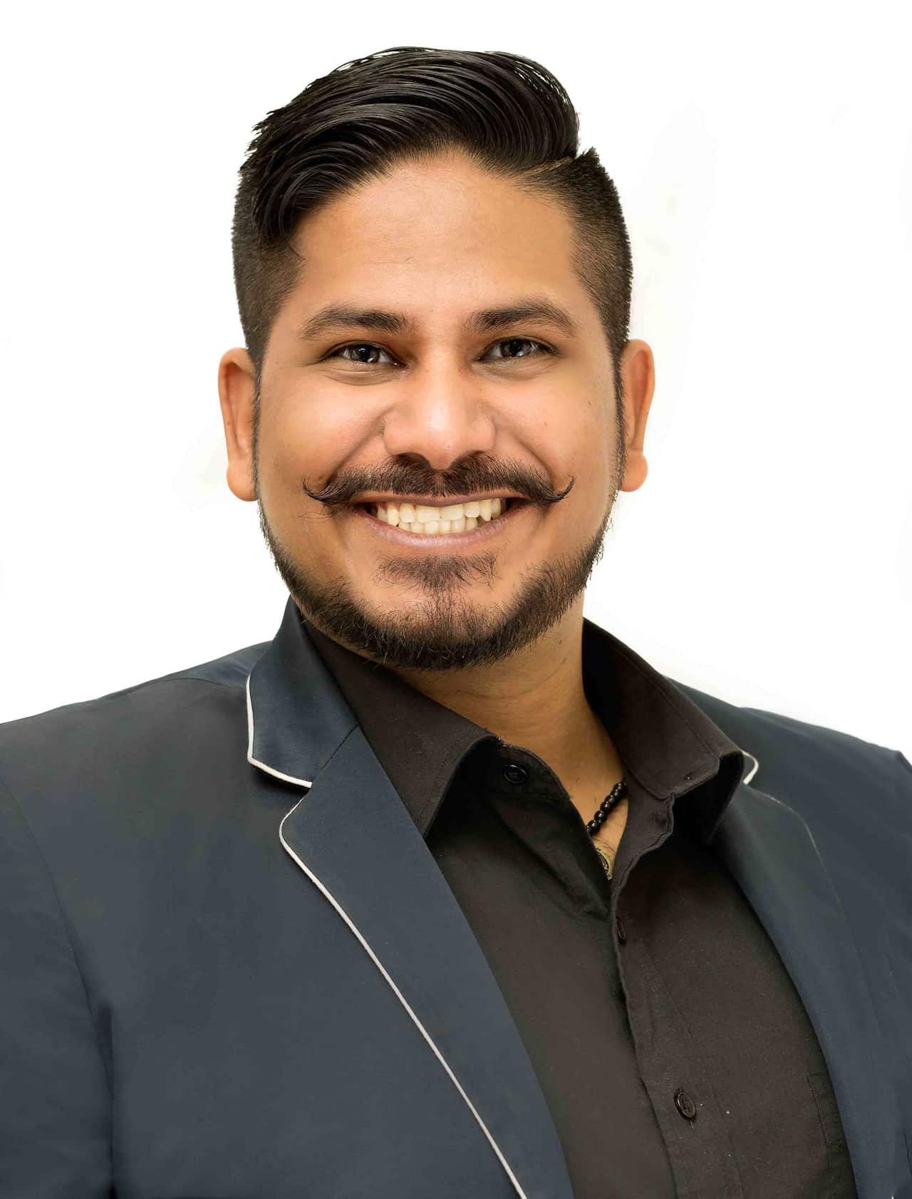 A portrait shot of a smiling man with a beard and moustache, wearing a dark blue jacket with light-coloured trim over a black collared shirt. He has styled dark hair and a beaded necklace is visible at his throat.