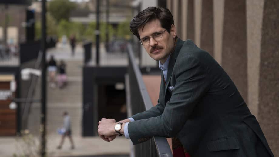 A man in a suit and wearing glasses leaning on a balcony looking towards the camera