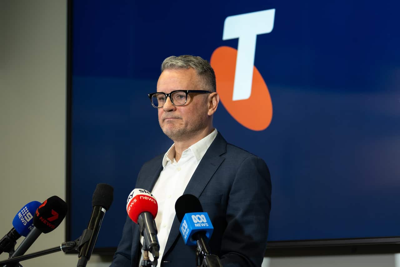 A middle aged white man in glasses and a suit stands in front of microphones and a Telstra logo.