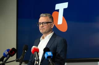 A middle aged white man in glasses and a suit stands in front of microphones and a Telstra logo.