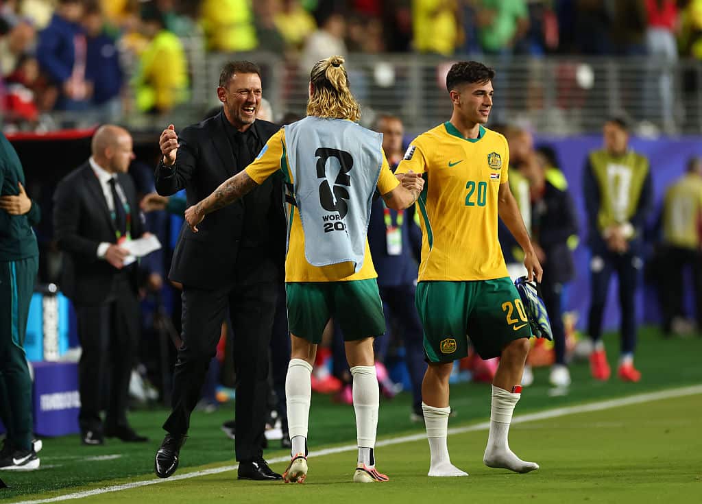A man in a black suit smiles and is about to hug a player wearing a grey bib facing him on the sideline. That player is shaking hands with another player in yellow shirt and green shorts facing the pitch.