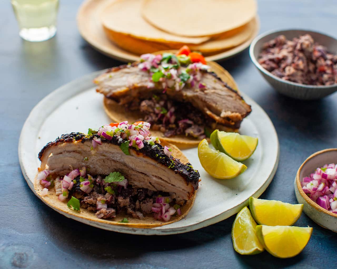 Two tortillas sit on a pale plate, each topped with a generous slice of cooked pork belly, topped with chopped red onion salsa. 