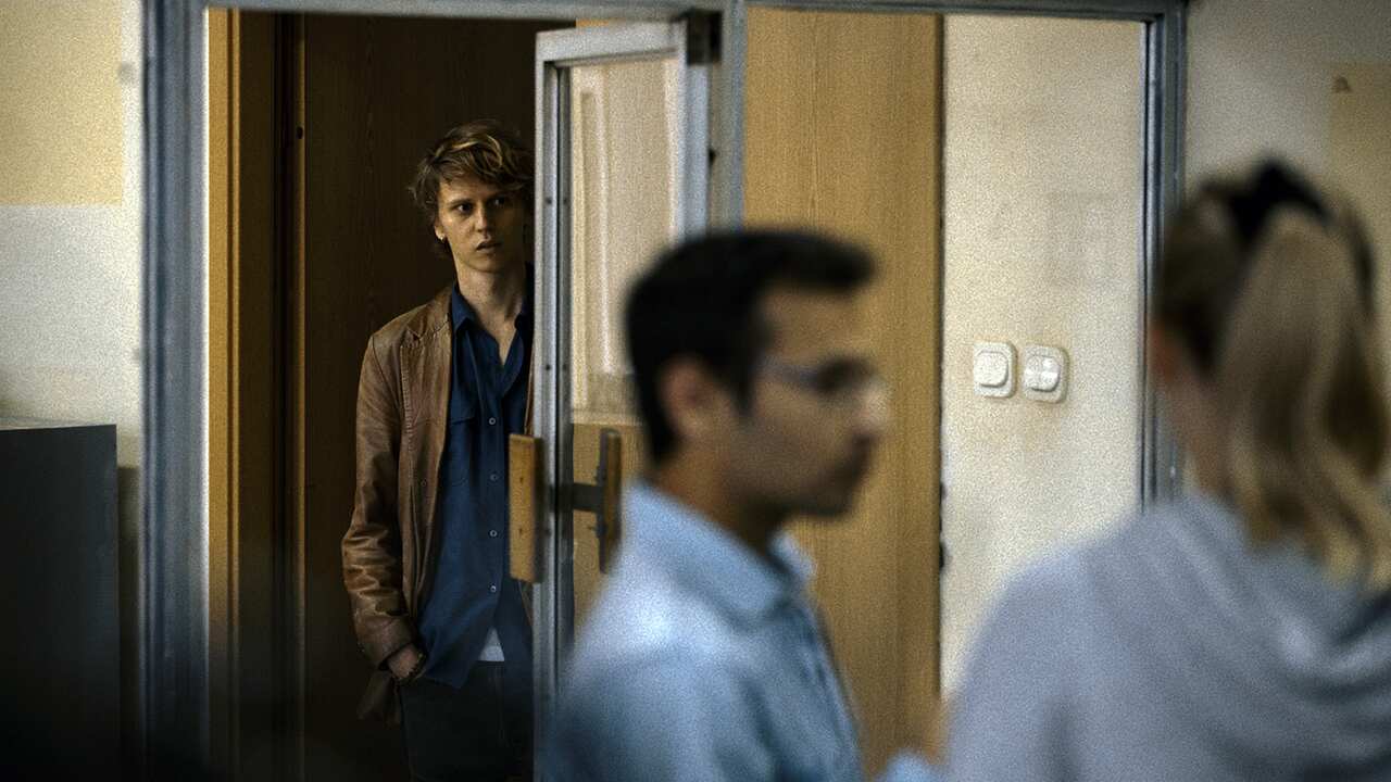 A young man stands in a doorway, looking into a room where several people can be seen standing. He has a slightly hesitant expression. 