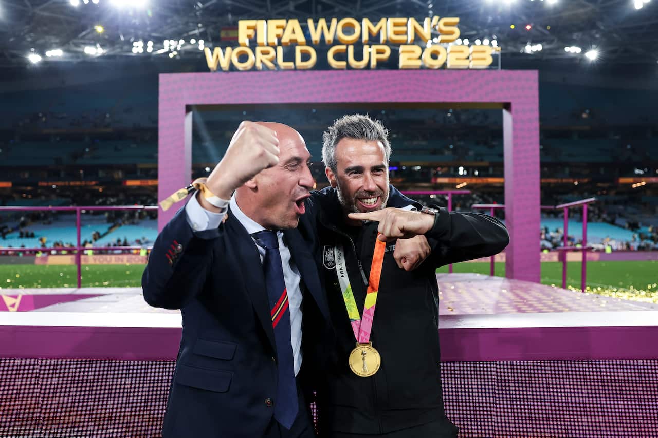 Luis Rubiales (left) wearing a suit and tie and Jorge Vilda (right), wearing a black tracksuit, celebrate in front of small purple stage on a football field. Vilda is wearing a gold medal around his neck.