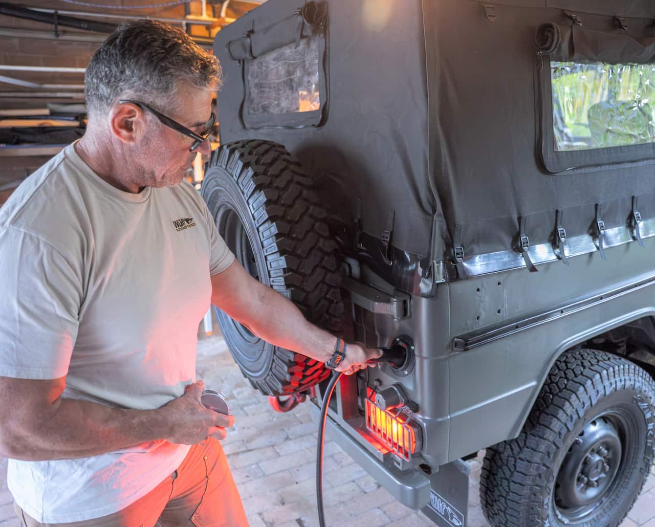 A man wearing a t-shirt stands beside an electric vehicle with the charger plugged in.