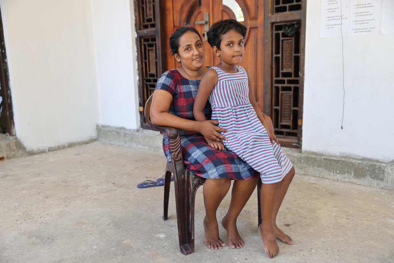 A young girl sits on her mother's lap on a chair in front of their house