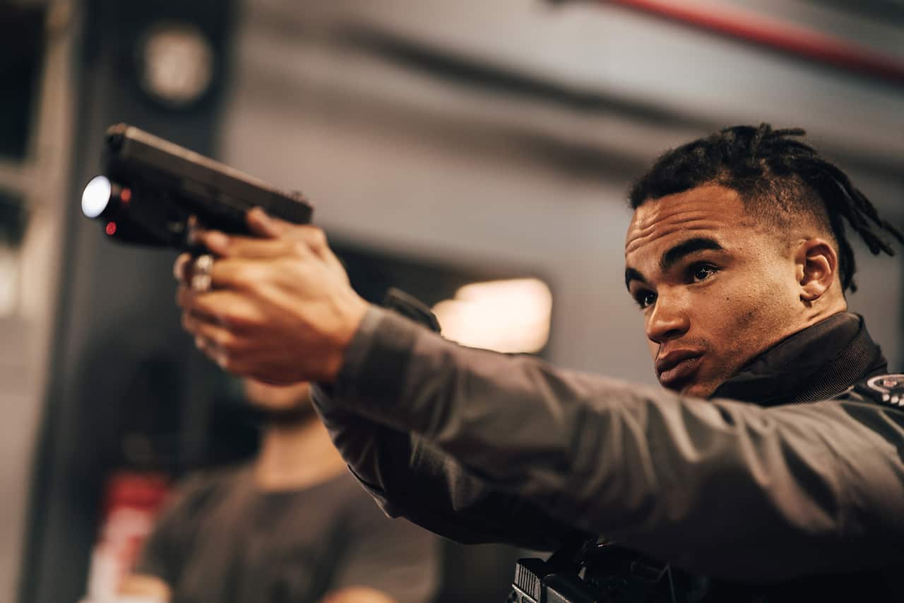 A man in a dark coat is seen from the waist up, pointing a gun to the left with both hands.