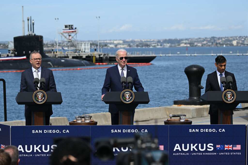 Three men standing at lecterns on a dock. There is a submarine in the water behind them