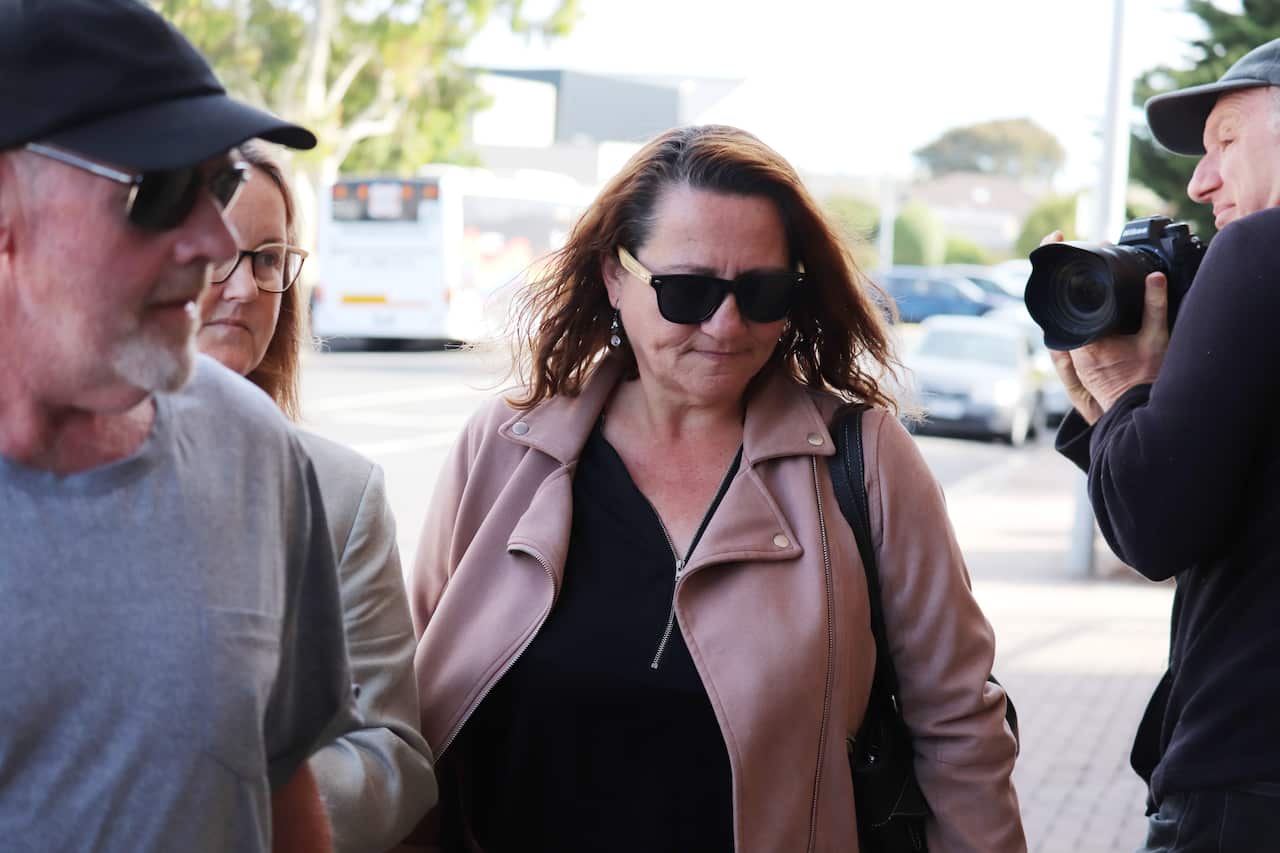 A woman wearing sunnies and a light pink jacket over a black shirt is walking outside.
