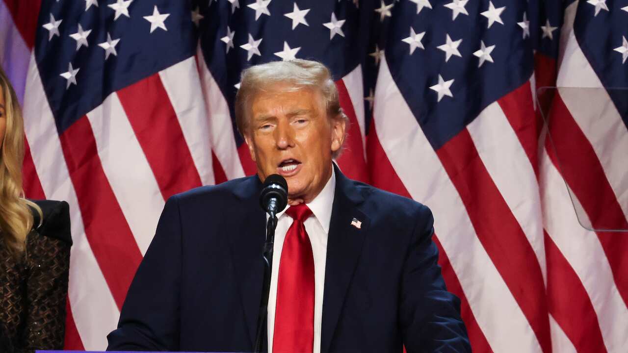 Donald Trump speaking into a microphone, with a large American flag behind him.