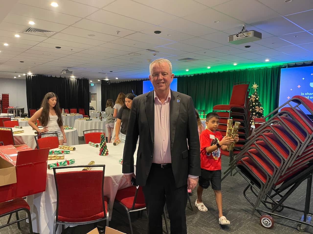 Man standing in front of some tables with Christmas decorations on them and kids around setting up.