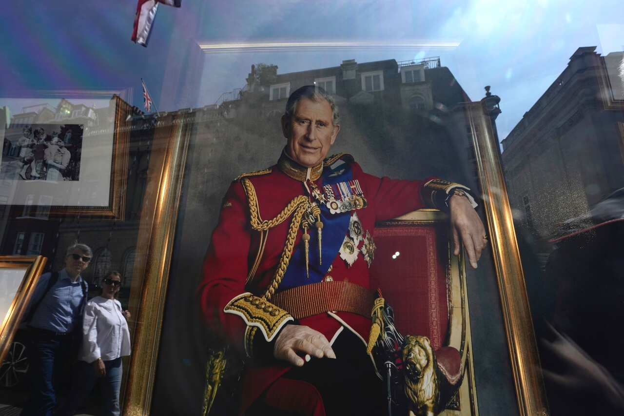 A man and a woman's reflections are seen in the window of a store selling King Charles' portraits.