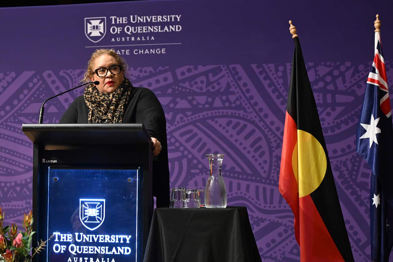 A woman in a black top and leopard-print scarf stands at a lectern with a University of Queensland logo on the front.