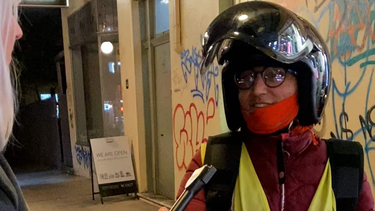 Female delivery rider wearing a black scooter helmet and a high visibility safety vest stands afront a graffitied wall. 