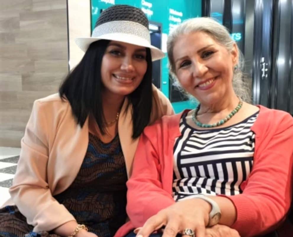 A woman in a white sun visor sits next to another woman in a coral cardigan.