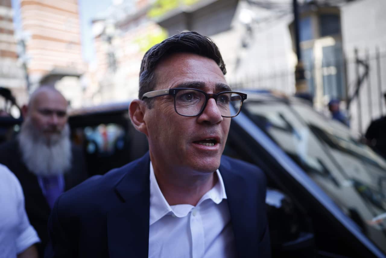A man wearing glasses, a black blazer and white shirt, stepping out of a black car.