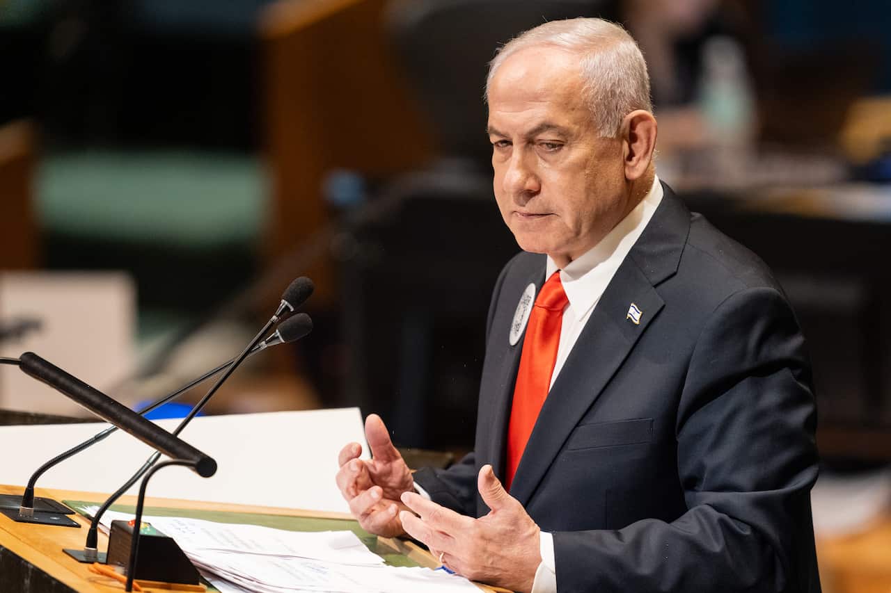A close-up photograph shows Benjamin Netanyahu, dressed in a dark suit and a bright red tie, speaking at a podium with two microphones in front of him. He is mid-gesture, looking intently to his left, with a serious expression on his face in a dimly lit, official setting.