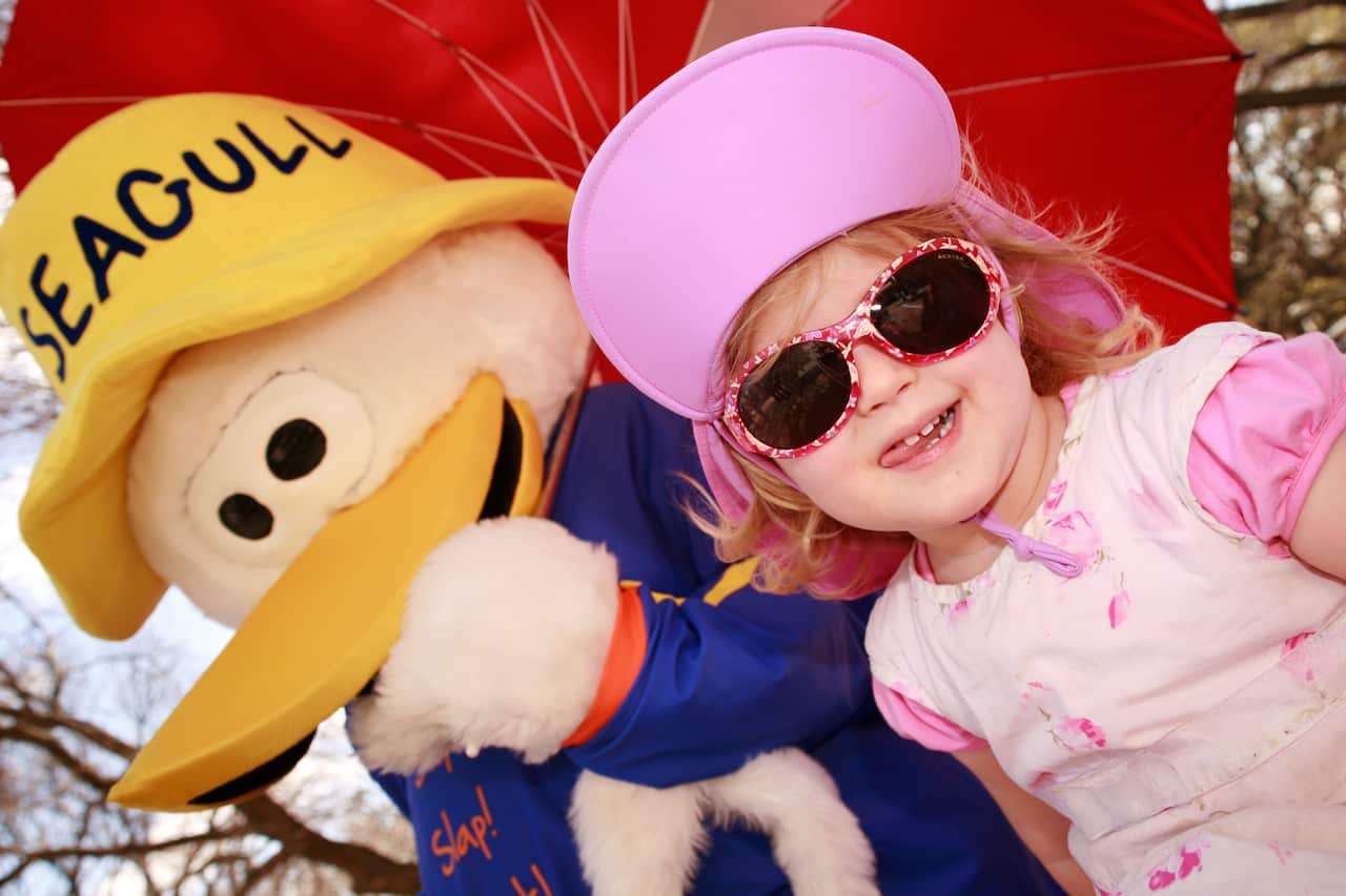 A little girl wearing a pink dress, hat, and sunglasses stands next to Sid the Seagull.