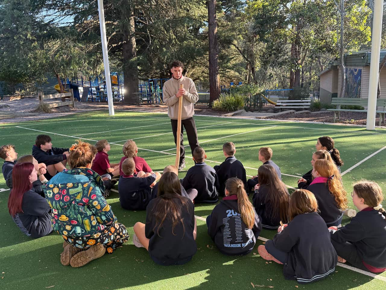 Young students sit outside on a basketball court as a man undertakes a cultural heritage workshop.