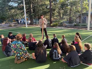 Young students sit outside on a basketball court as a man undertakes a cultural heritage workshop.