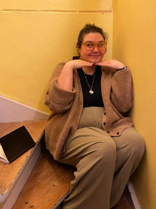 A person smiling while sitting on stairs. Their hands are under their chin, framing their face.