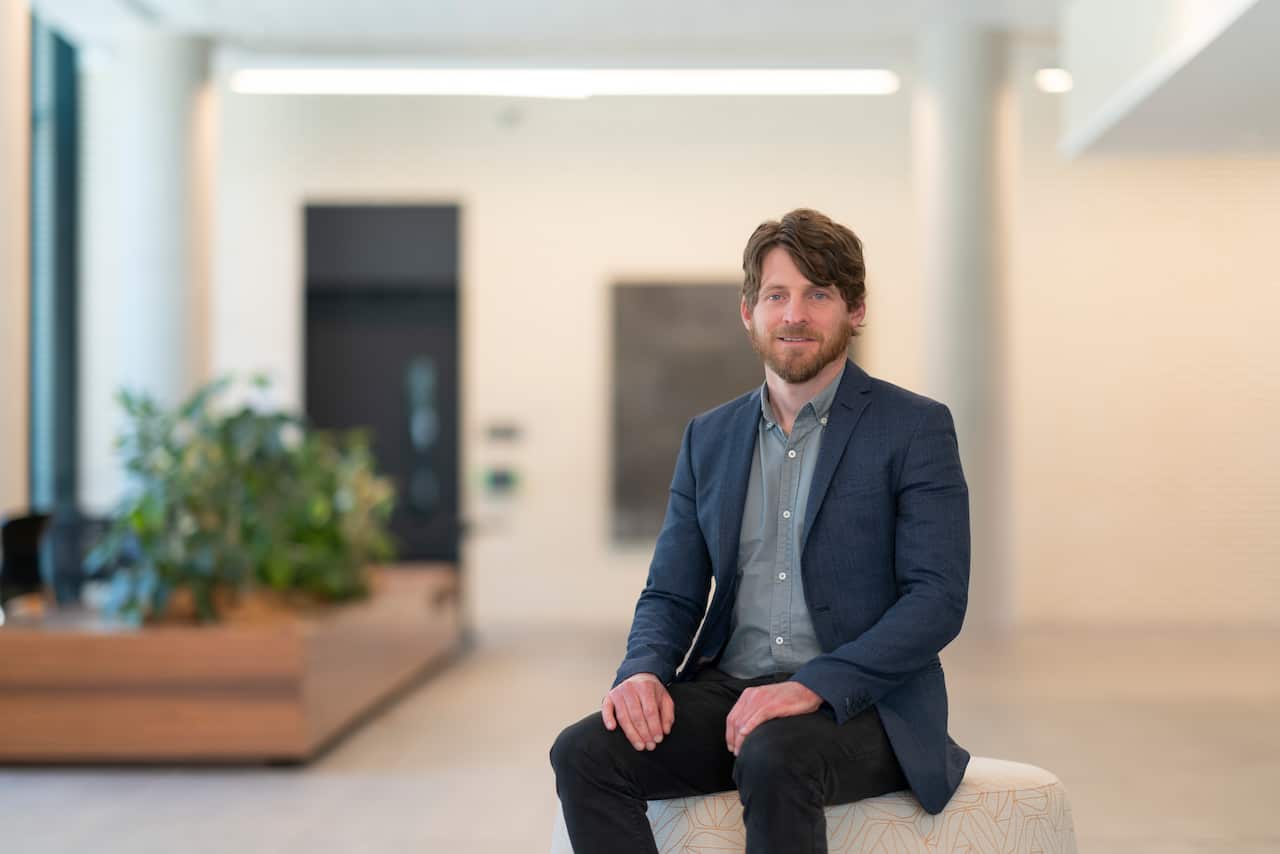 A man wearing a blue blazer over a grey shirt sits on a pouf stool, smiling at the camera.