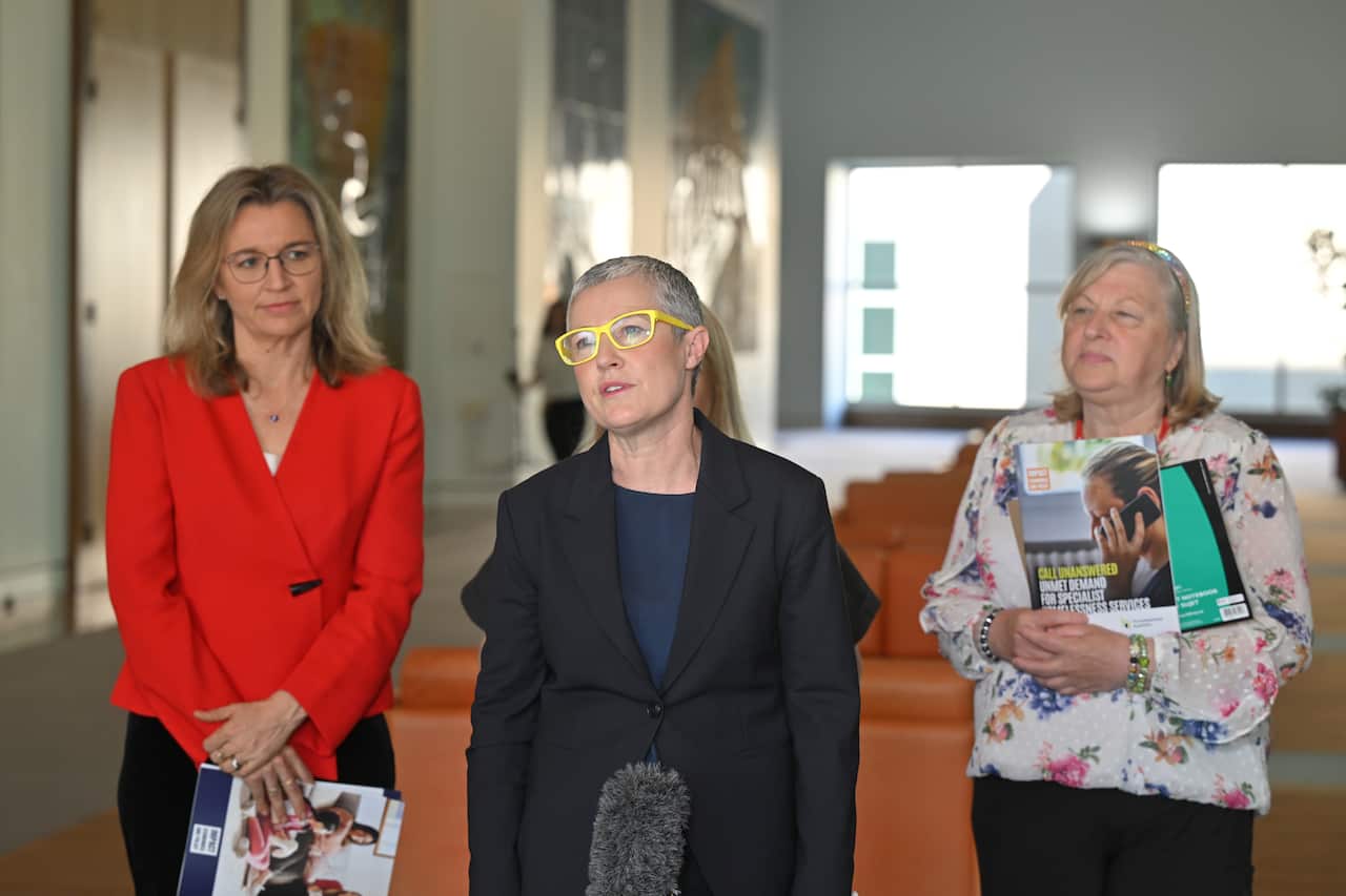 A woman with yellow glasses frames speaks into microphone, two women stand beside her.
