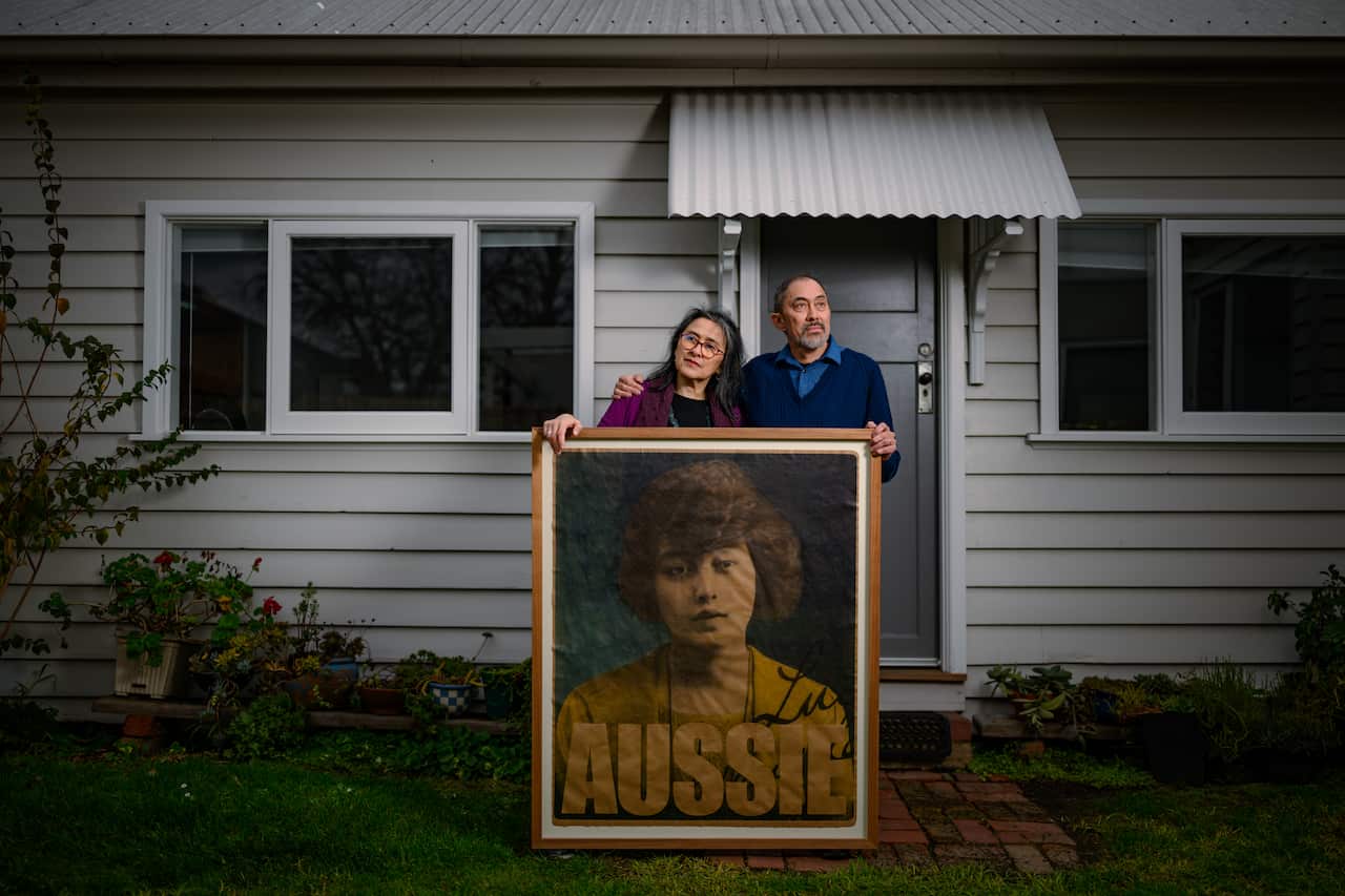A brother and sister stand outside a grey-clad home holding a large, framed poster of a woman with the text 'Aussie'. They have their arms wrapped around each other and look off into the distance.