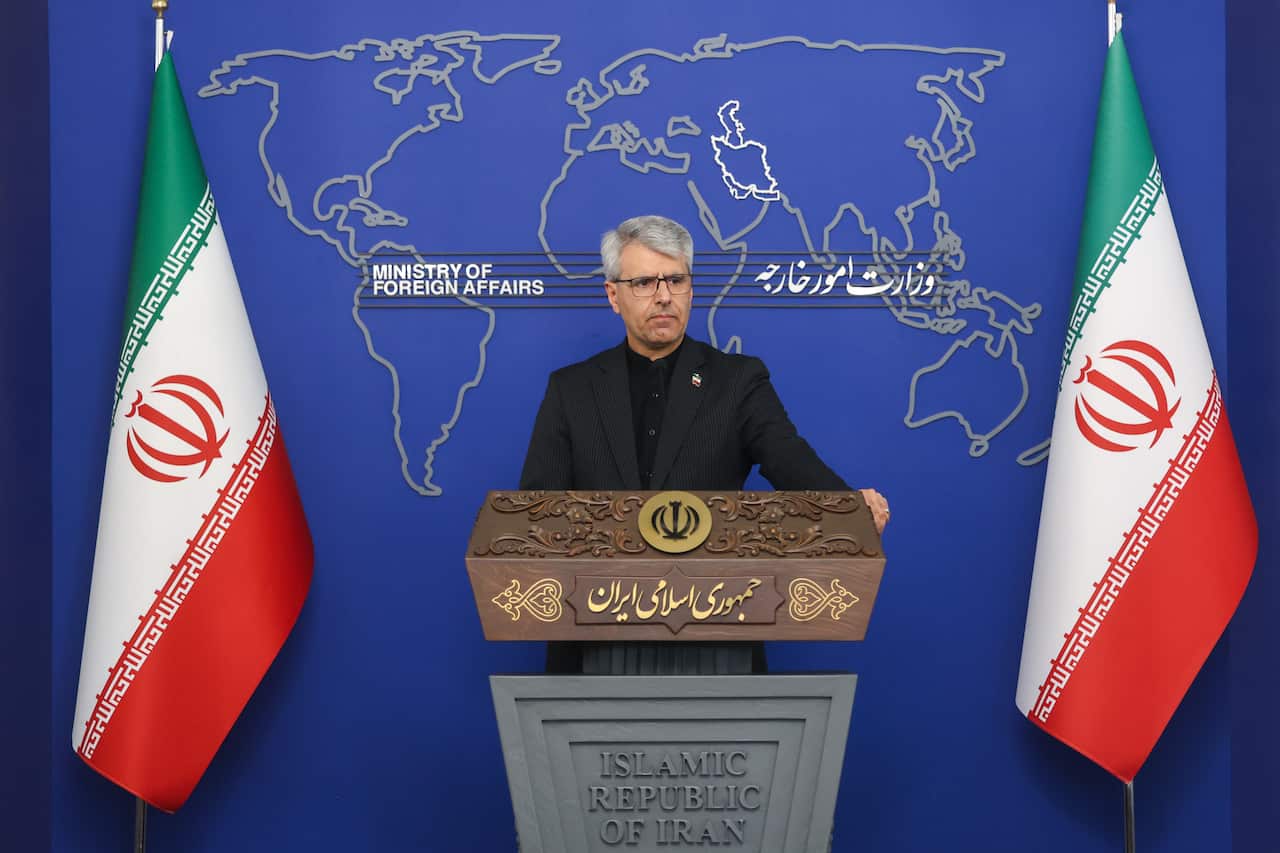 A man with grey hair in black suit stands behind a lectern, in between two Iranian flags.