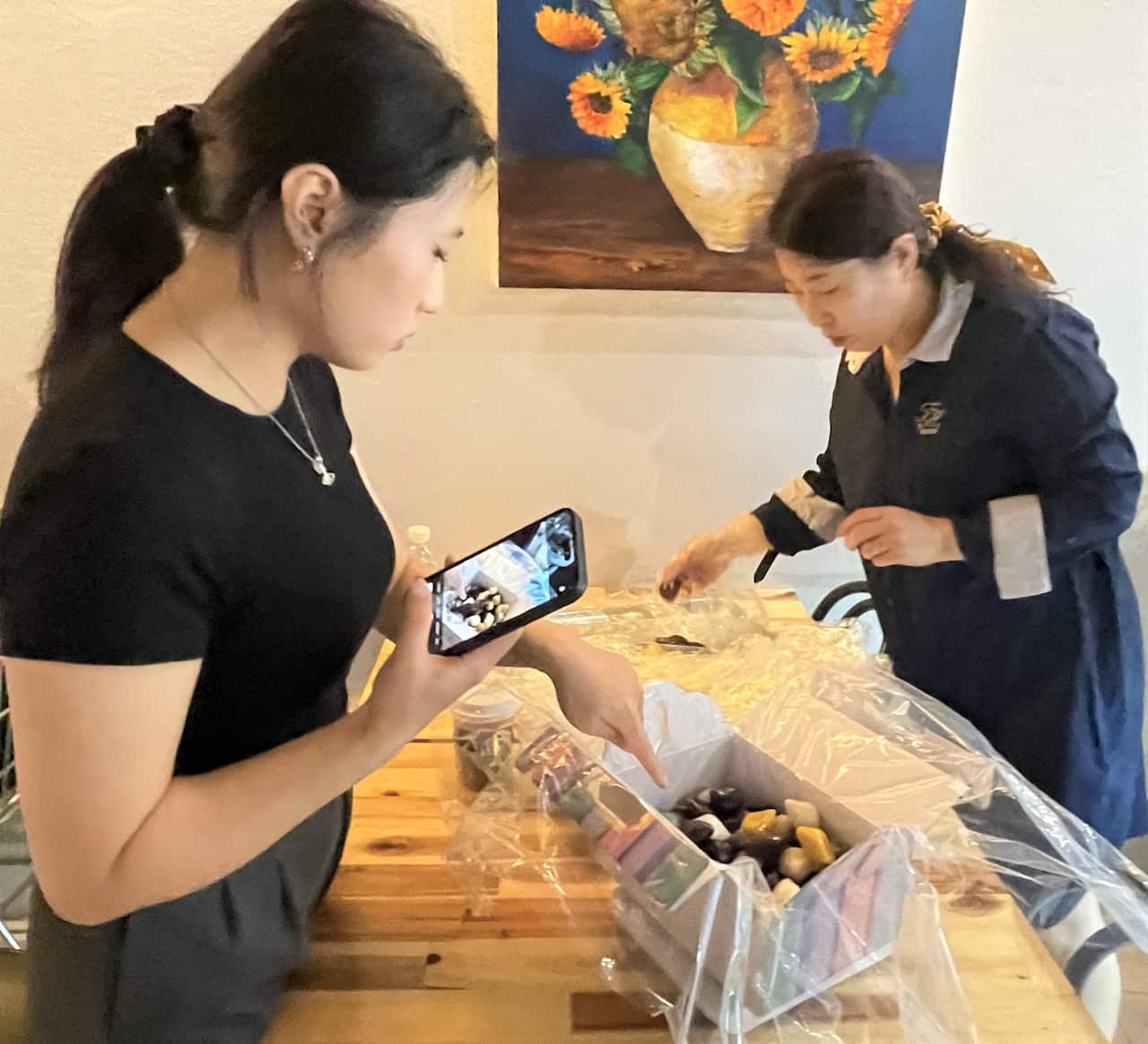 A woman in a black t-shirt standing behind a counter. A woman in a dark dress with white cuffs is on the opposite side of the counter to her left. There is a box of rice cakes on the counter.