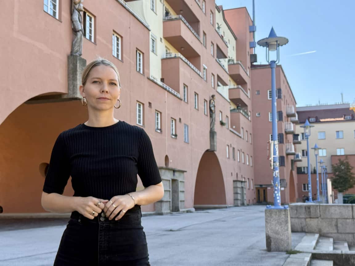 A blonde woman stands in front of a large apartment complex with metallic statues adorning the walls. 