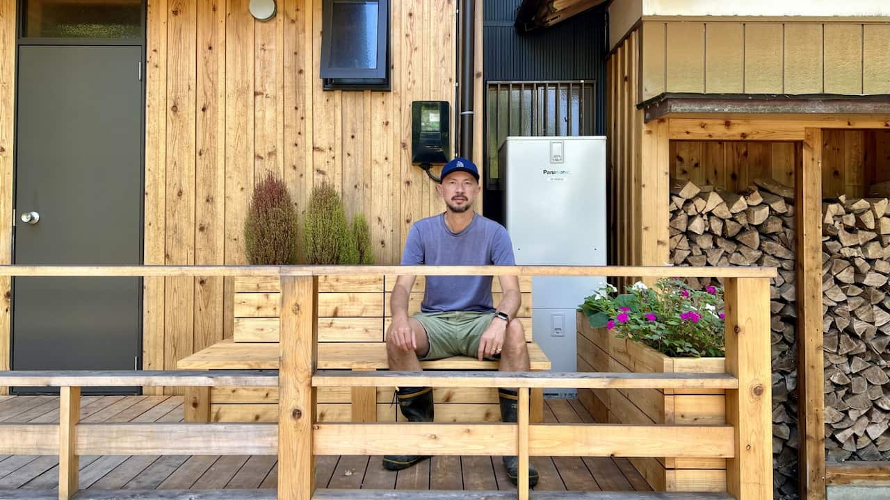 A man wearing a grey t-shirt, khaki shorts and a baseball cap sitting on a wooden bench on a timber-clad veranda