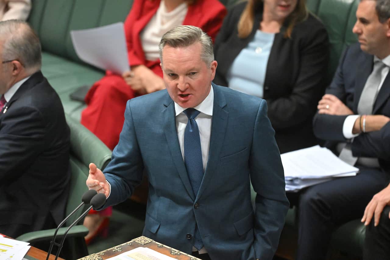 A man in a blue suit and blue tie and short white hair is speaking, with people in business suits sitting behind him in the parliament.