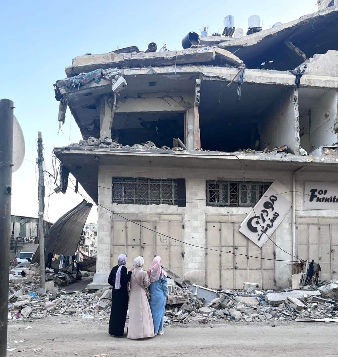 Three women stand in front of a destroyed home. On the floor beside them is rubble. 