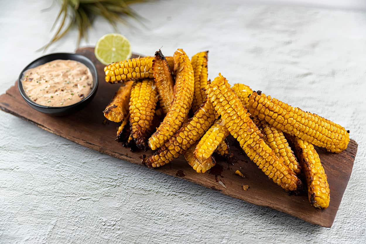 Long pieces of cooked corn sit on a long wooden board, along with a bowl of sauce and a piece of lime. 