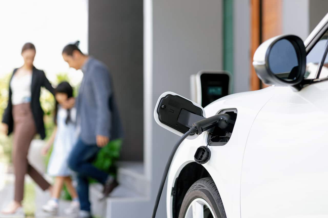 A cord attached to an electric vehicle charging it in the foreground and a blurred family in the background.