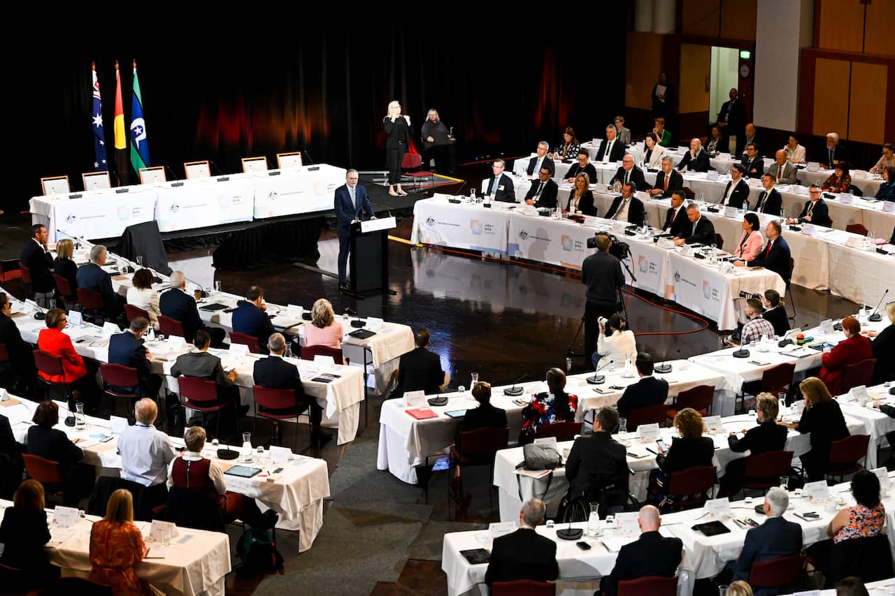 A man standing at a podium. Around him are people sitting at tables.