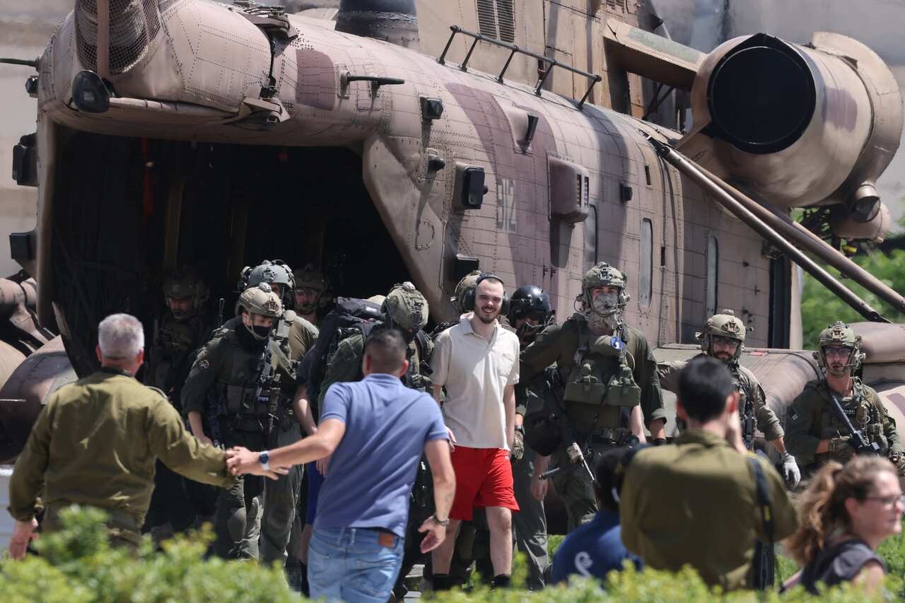 A man wearing headphones and red shorts smiles as soldiers escort him from a military helicopter.
