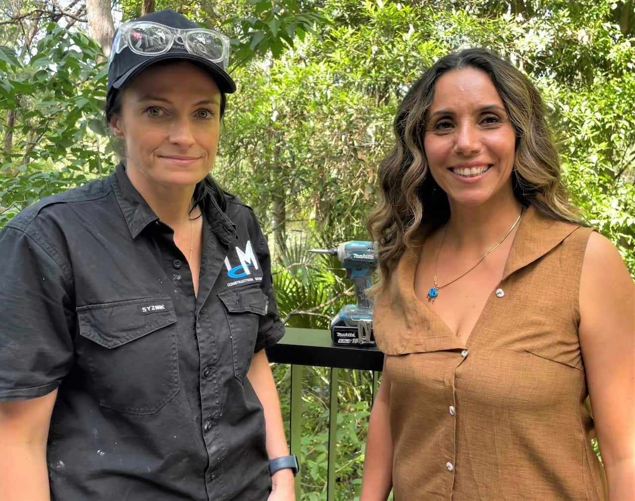 Two women standing on a balcony with a bush backdrop.