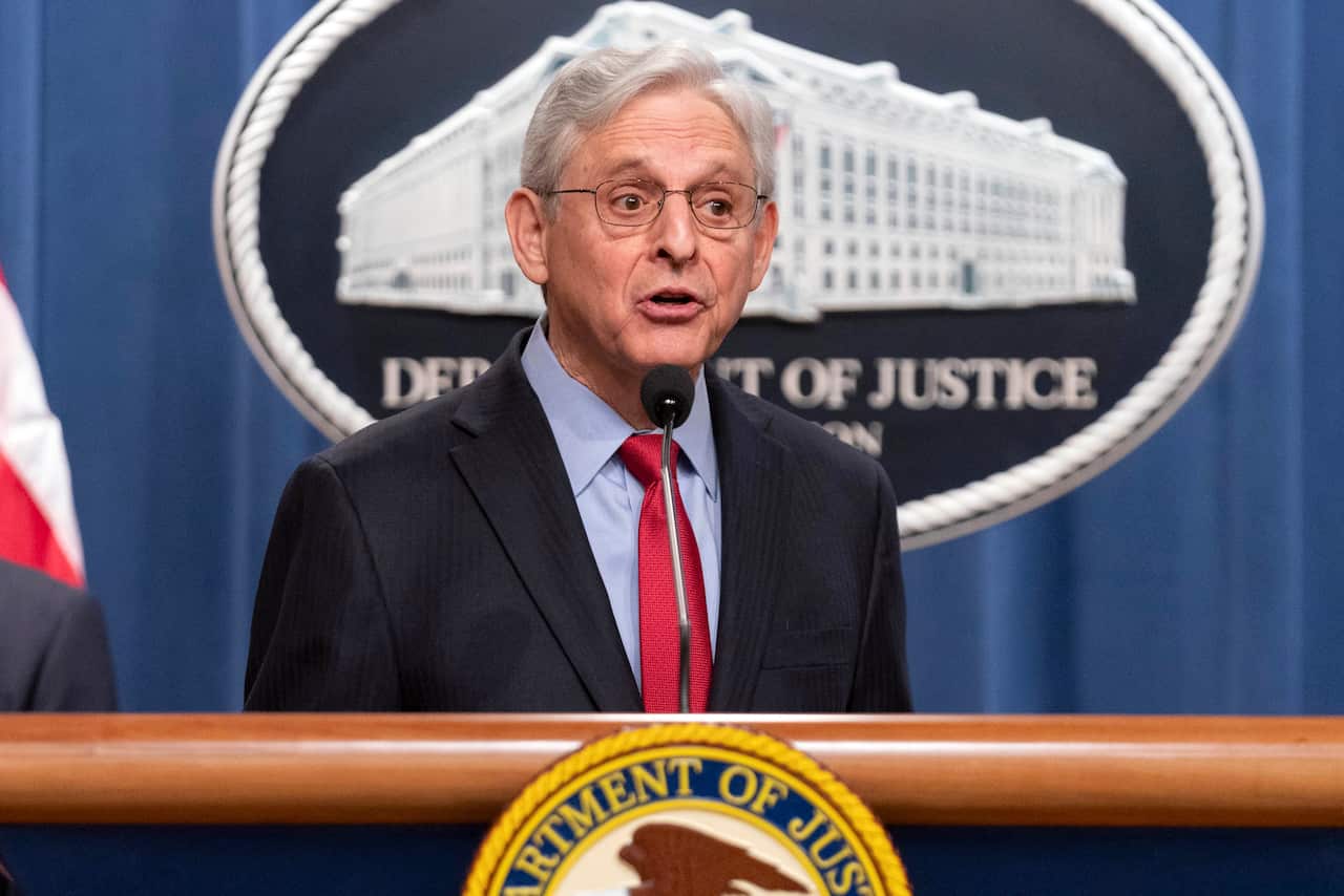 A man wearing a suit and tie speaking into a microphone at a podium. The Department of Justice logo is behind him.