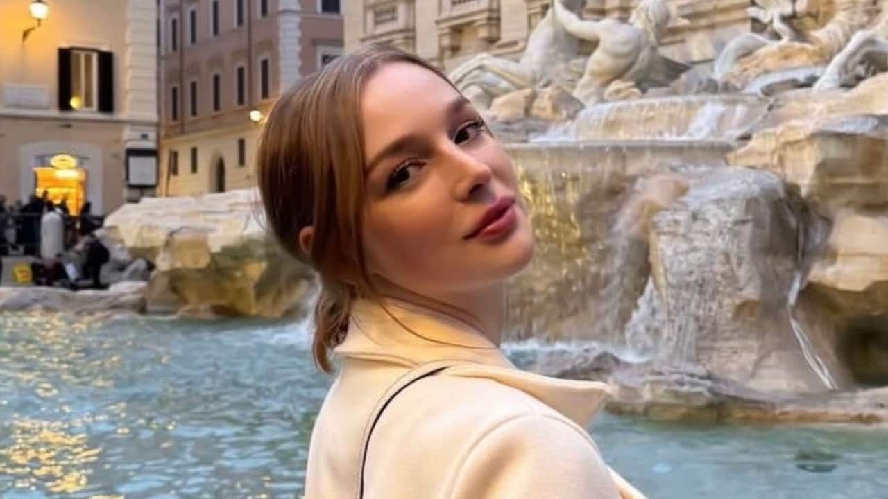 a young woman with red hair in a white coat poses in front of the trevi fountain