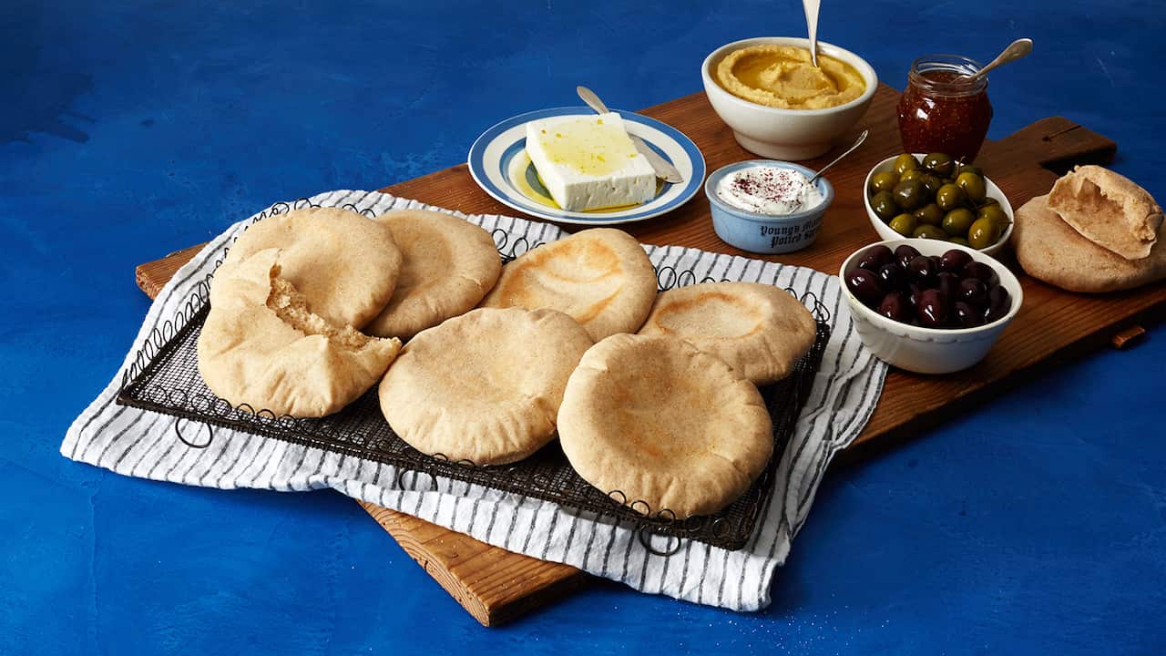 Seven puffy pita breads, one of them torn open, sit on a decorative black metal tray, on top of a striped tea towel. The tray and towel sit on a wooden board that also has olives, cheese and dips, along with more pita breads. 